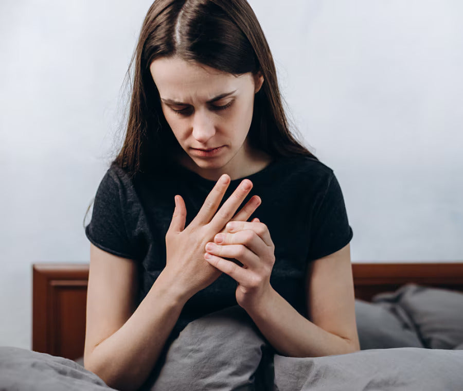 Mujer sentada en su cama, con expresión de dolor, sujetándose las manos entumecidas y doloridas, síntoma común de artritis o rigidez matutina en las articulaciones.